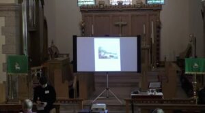 Church interior with a presentation screen displaying an image, speaker preparing notes at podium.