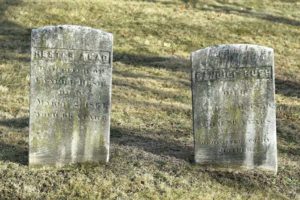 Two aged tombstones on a grassy cemetery, casting soft shadows in the sunlight.