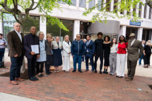 Group of people posing outdoors in business attire at an event in front of a building with trees and bricks.