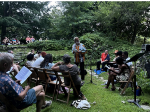 Outdoor gathering with people seated, listening to a speaker in a lush garden setting.