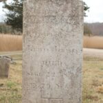 Old weathered gravestone in a cemetery, surrounded by grass and trees in the background.
