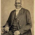 Vintage portrait of a distinguished elderly man in a formal suit, sitting, with a neutral background.