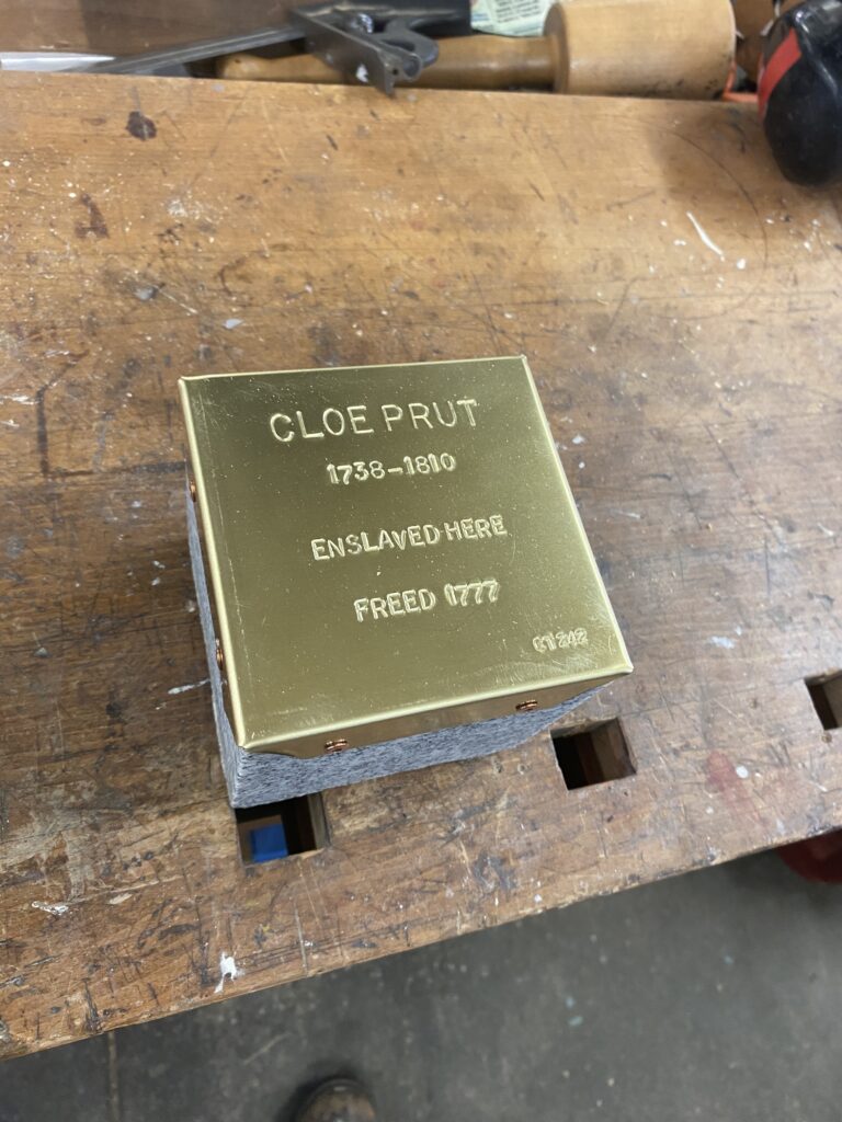 Engraved brass plaque reading Cloe Prut, 1738-1810, Enslaved here, Freed 1777 on a wooden workbench.
