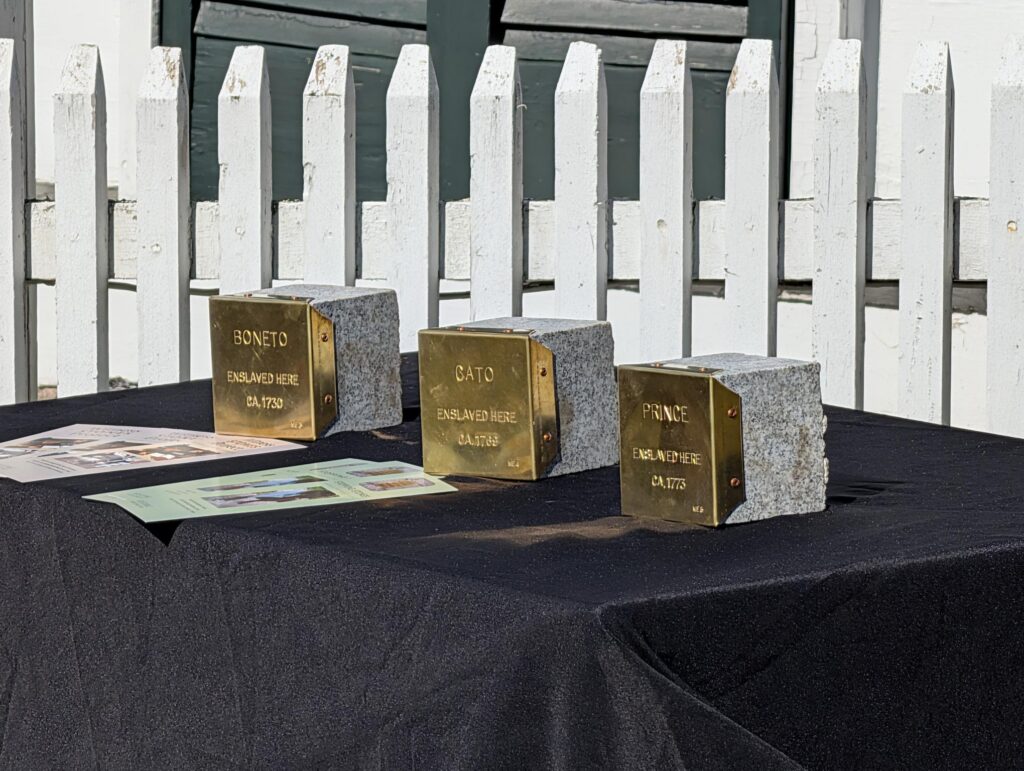 Memorial stones honoring enslaved individuals Boneto, Cato, and Prince displayed on a table in front of a white fence.