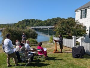 Outdoor speech near river with audience seated on grass, white building and bridge in background under clear blue sky.